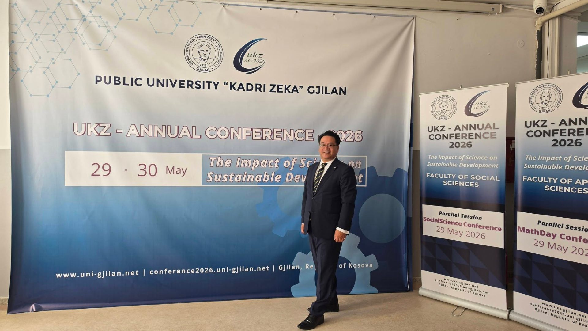 Dr. Glenn Agung Hole in front of the UKZ Annual Conference 2026 banner at Public University Kadri Zeka, Gjilan